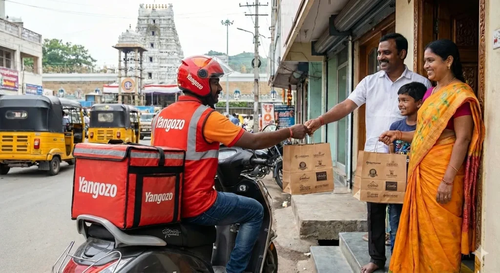 A Yangozo delivery partner in a red branded uniform handing over bags of fresh food to a smiling family at their doorstep, with a scenic view of a Tirupati temple in the background.